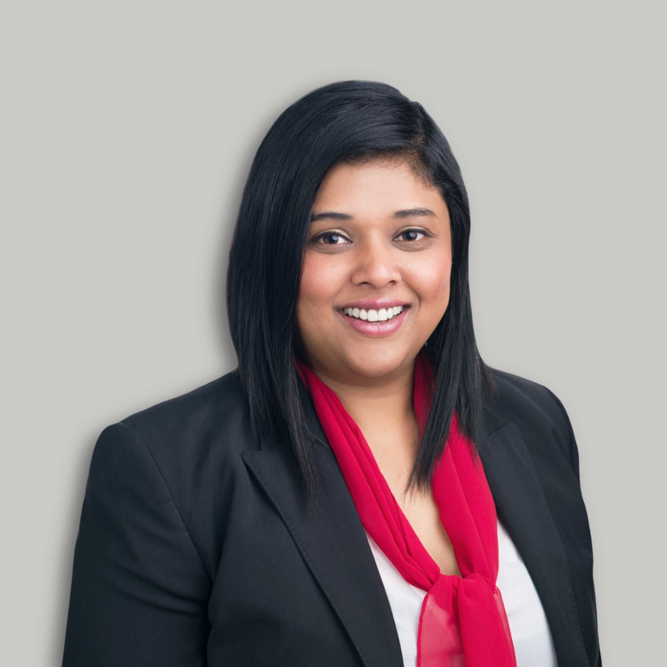 Priya Ravi smiles at the camera, wearing a black blazer and red scarf.